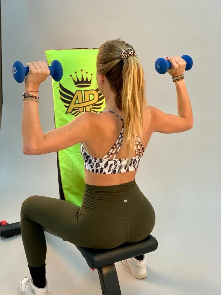 Chica entrenando hombro con mancuernas y la toalla verde flúor sujeta al respaldo